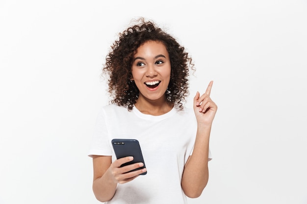 Portrait of a pretty cheerful casual african girl standing isolated over white wall, using mobile phone, pointing finger away