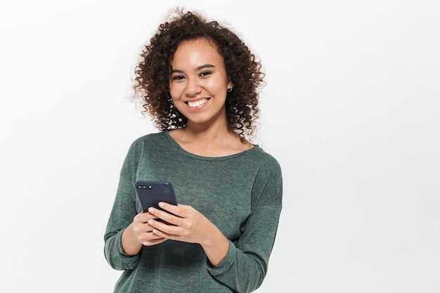 Portrait of a pretty cheerful casual african girl standing isolated over white wall, using mobile phone