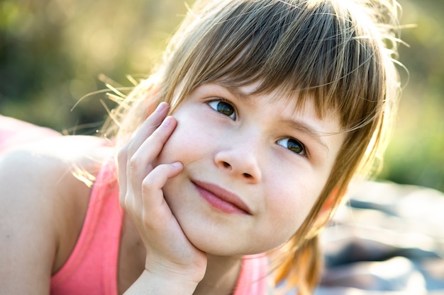 Portrait of pretty child girl with gray eyes and long fair hair leaning on her hands smiling happily outdoors. Cute female kid on warm summer day outside.