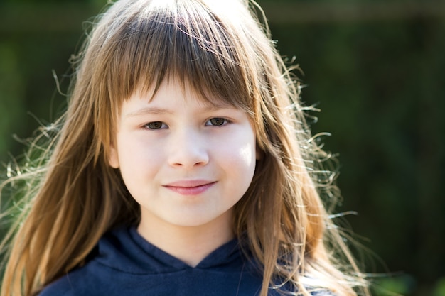 Portrait of pretty child girl with gray eyes and long fair hair smiling outdoors on blurred bright background. Cute female kid on warm summer day outside.