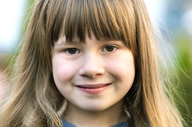 Portrait of pretty child girl with gray eyes and long fair hair smiling outdoors on blurred green bright background. Cute female kid on warm summer day outside.