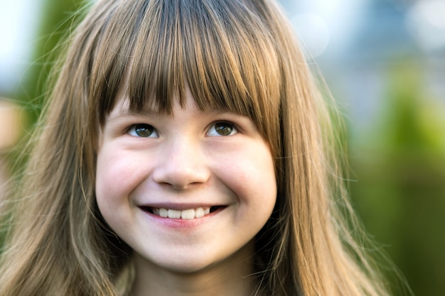 Portrait of pretty child girl with gray eyes and long fair hair smiling outdoors on blurred green bright background. Cute female kid on warm summer day outside.