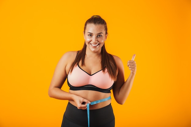 Portrait of pretty chubby woman in sportive bra measuring her waist with meter, isolated over yellow background