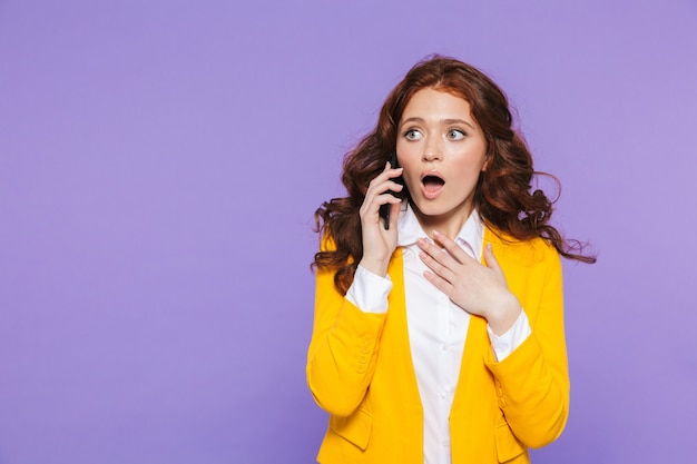 Portrait of a pretty confused young redheaded woman standing over violet, using mobile phone