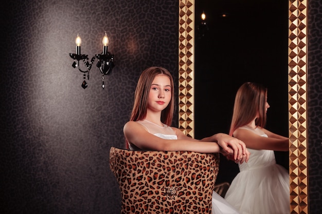 Portrait of pretty cute teenager girl in an elegant dress at mirror in dark stylish interior of living room. Children emotions and posing. Concept of style, fashion and beauty. Copy space for site