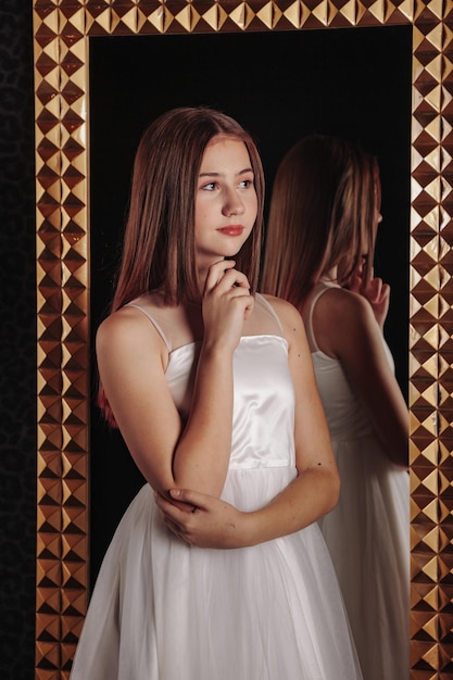 Portrait of pretty cute teenager girl in an elegant dress at mirror in dark stylish interior of living room Children emotions and posing Concept of style fashion and beauty Copy space for site