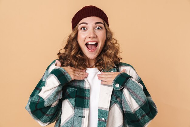 Portrait of pretty excited woman in knit hat expressing surprise and smiling isolated over beige wall