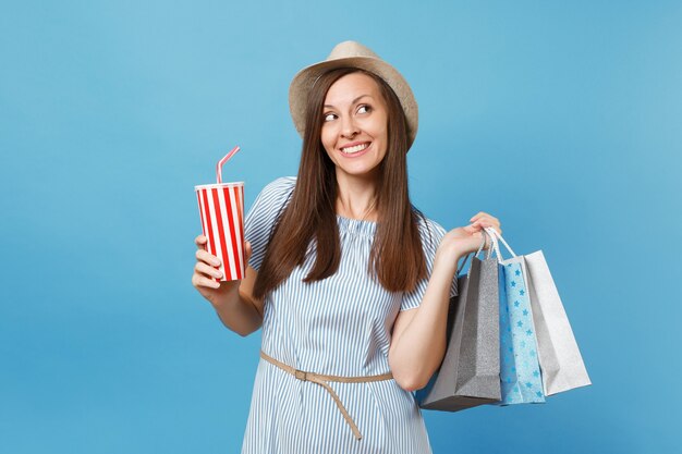 Portrait pretty fashionable smiling girl in summer dress, straw hat holding packages bags with purchases after shopping, soda water cup isolated on blue pastel background. copy space for advertisement