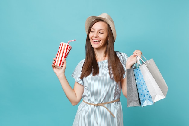 Portrait pretty fashionable smiling girl in summer dress, straw hat holding packages bags with purchases after shopping, soda water cup isolated on blue pastel background. Copy space for advertisement