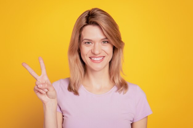 Portrait of pretty friendly lady toothy smile show v-sign look camera on yellow background