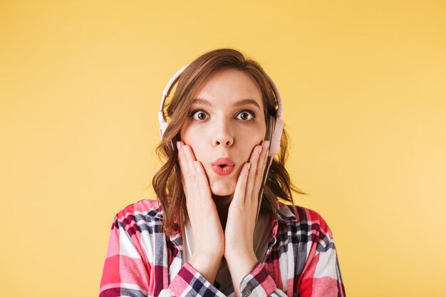 Portrait of pretty girl in colorful shirt standing in headphones and amazedly looking in camera on over pink background