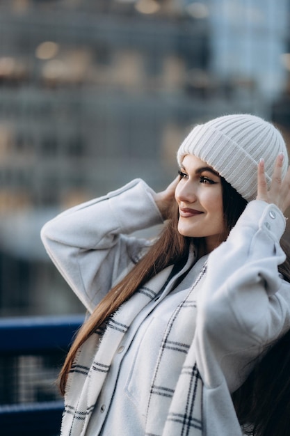 Portrait of pretty girl dreaming smiling on the city background Side view of beautiful girl dressed in white hat and checkered scarf holding her hat