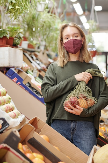 Portrait of pretty girl in mask holding net bag of organic products at farmers market during coronavirus