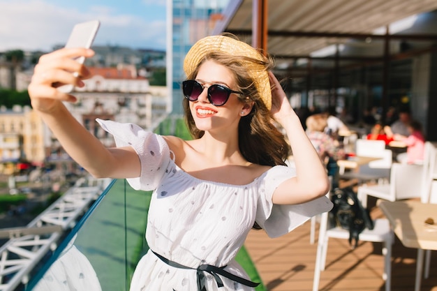 Portrait of pretty girl with long hair in sunglasses standing on the terrace. She wears a white dress with naked shoulders, red lipstick and hat . She is making selfie on phone.