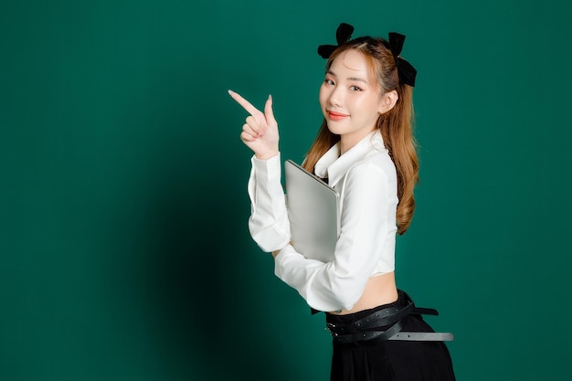 Portrait of pretty joyful teenage asian student woman in white shirt and black short skirt with slim body holding laptop pointing to copy space on isolated green background mock up copy space