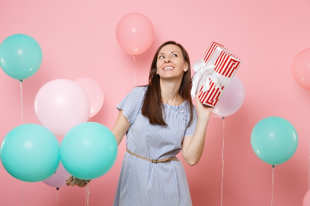 Portrait of pretty joyful woman wearing blue dress dreaming looking up holding red box with gift present and colorful air balloons on bright trending pink background. Birthday holiday party concept.