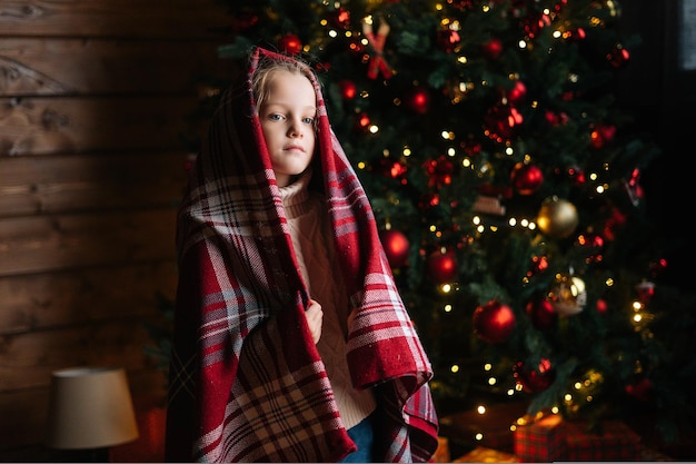 Portrait of pretty little blonde curly child girl wrapped in plaid looking at camera standing by