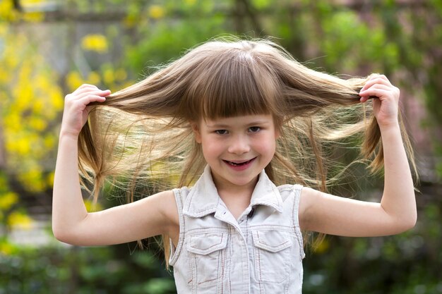 Portrait of a pretty little long-haired blond preschool girl in sleeveless white dress smiling shyly into camera against blurred outdoors background. Innocent happy childhood concept.