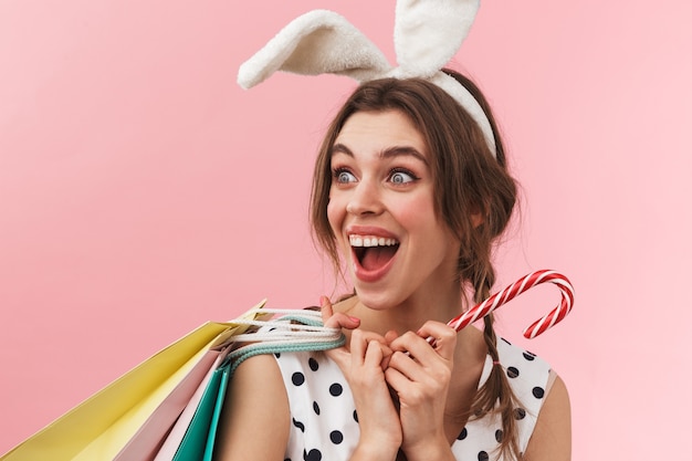 Portrait of a pretty lovely girl wearing bunny ears standing isolated, carrying shopping bags, holding candy cane