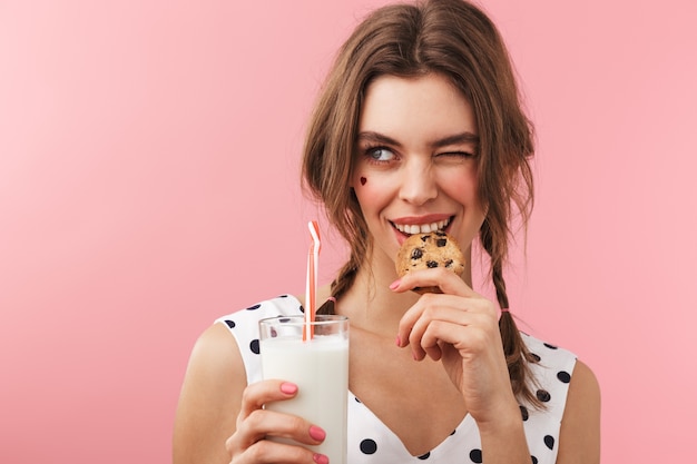 Portrait of a pretty lovely girl wearing dress standing isolated, eating chocolate chip cookie, drinking milk