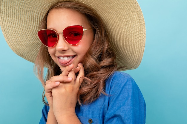 Portrait of pretty model girl with wavy chestnut hair in dress, big hat, red sunglasses smiles isolated on blue