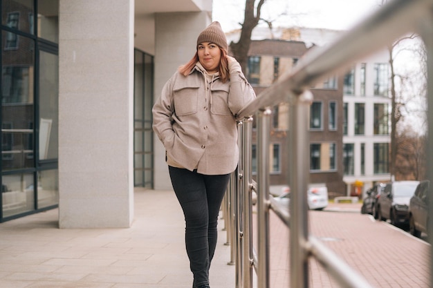 Portrait of pretty overweight woman in warm hat and jacket standing posing near railing of office building at city street in cloudy autumn day