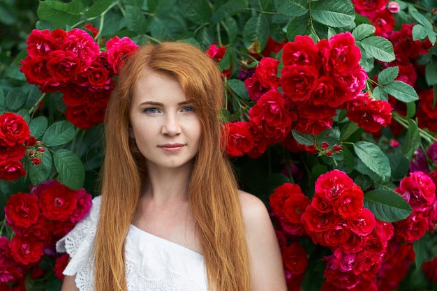 Portrait of a pretty redhead girl dressed in a white light dress, blooming roses. Outdoor.