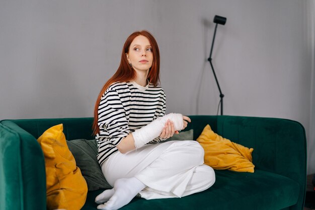 Portrait of pretty redhead young woman with broken right hand wrapped in white plaster bandage sitting on sofa pensive looking away