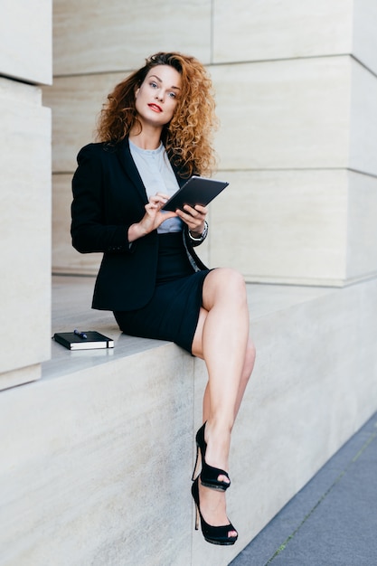Portrait of pretty slim woman with curly hair, wearing black jacket, skirt and high-heeled shoes, using modern tablet computer