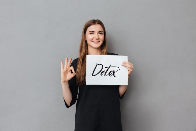 Portrait of pretty smiling lady standing and holding postcard while happily looking in camera on gray background