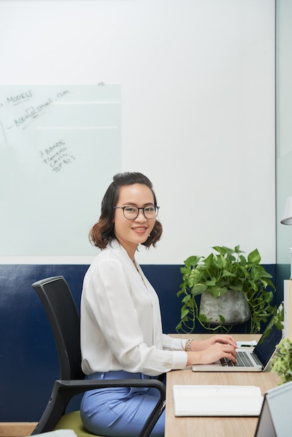 Portrait of pretty smiling young businesswoman in glasses working on laptop at office desk and looking at camera