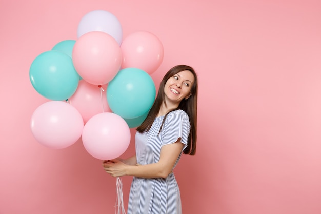 Portrait of pretty smiling young woman in blue dress holding colorful air balloons look aside on copy space isolated on bright pink background. Birthday holiday party, people sincere emotions concept.