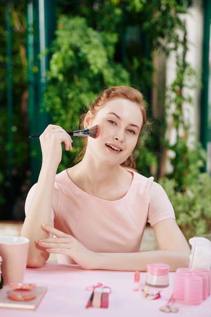 Photo portrait of pretty smiling young woman sitting at vanity table and applying make-up when getting ready for event