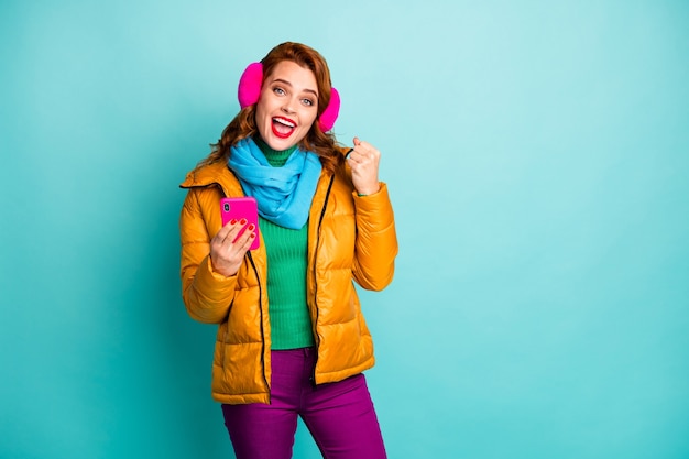 портрет красивой дамы путешествия держать телефон празднует достижение первого дохода онлайн деньги носить модные повседневные желтые пальто шарф фиолетовые брюки.