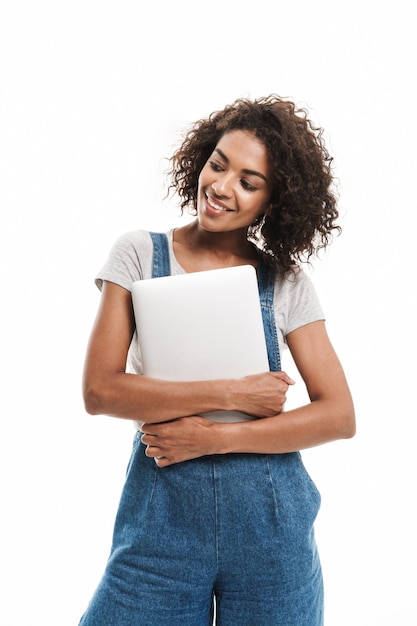 Portrait of pretty woman dressed in denim overalls smiling and holding laptop isolated over white wall