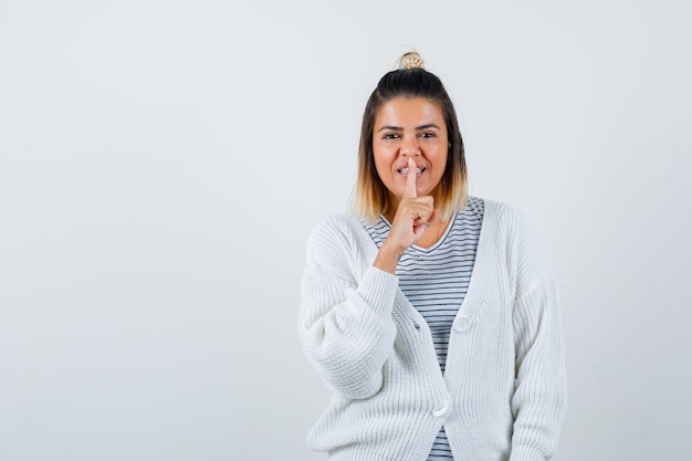 Portrait of pretty woman showing silence gesture in t-shirt, cardigan and looking cheerful