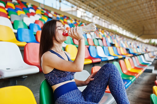 Photo portrait of a pretty woman in sportswear sitting and drinking water in the stadium.