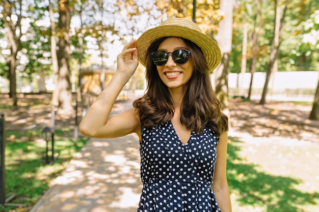 Portrait of pretty woman walking in the green summer park dressed in dress and hat. Holds her hat. Summer sunny day.