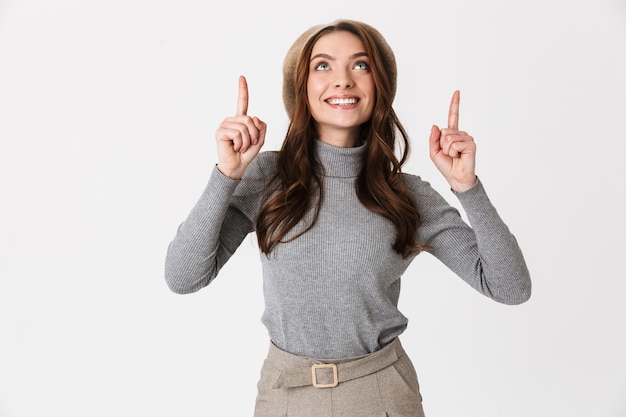 Portrait of pretty woman wearing hat smiling and pointing fingers upward at copyspace isolated over white wall