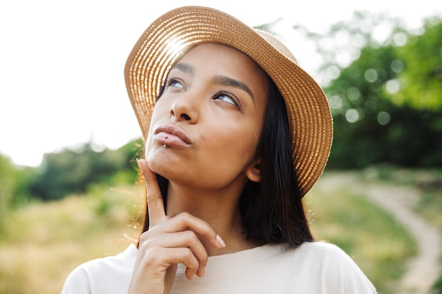 Portrait of pretty woman wearing straw hat and lip piercing looking upward while walking in green park