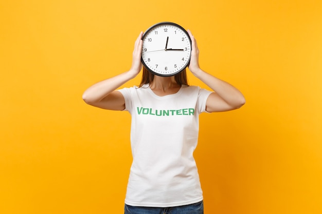 Portrait of pretty woman in white t-shirt with written inscription green title volunteer hold round clock isolated on yellow background. voluntary free assistance help, charity grace work time concept