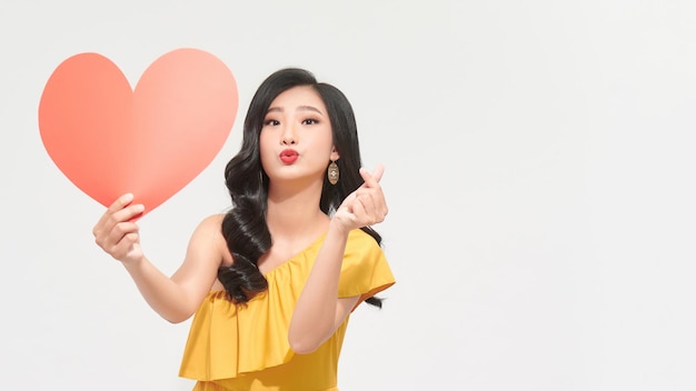 Portrait pretty woman in yellow dress sends air kiss with red heart over white background