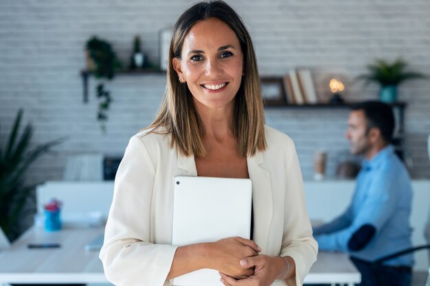 Portrait of pretty young businesswoman looking at camera while holding digital tablet. In the background, her colleagues working in the office.