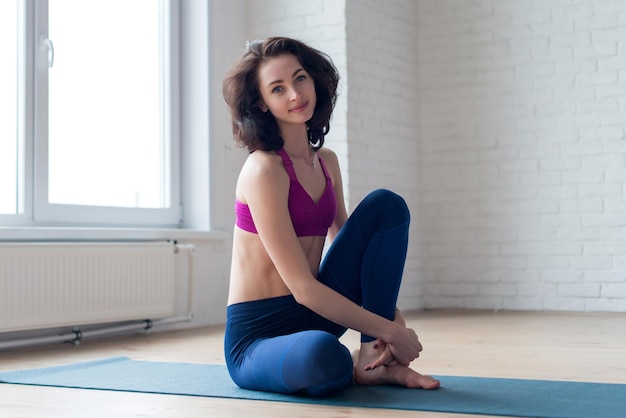 Portrait of pretty young Caucasian sportswoman with dark hair wearing sports bra and leggings sitting on mat barefoot smiling and looking at camera