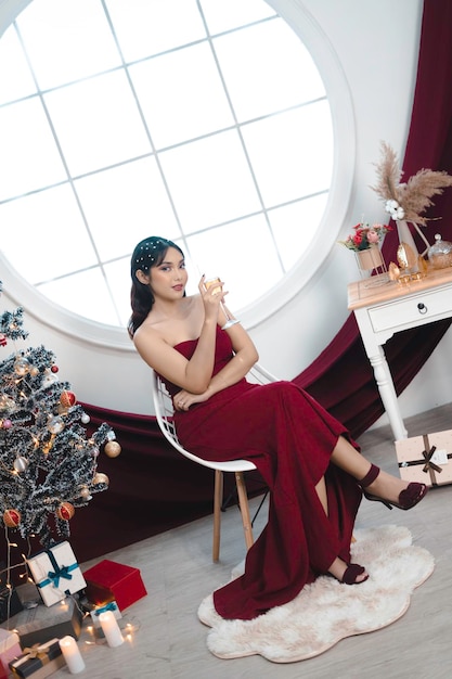 Portrait of pretty young girl cozy sit down and drinking wine wear red gown in decorated Christmas living room indoors