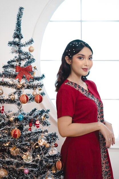 Portrait of pretty young girl decorating christmas tree smiling wearing red gown in decorated christmas living room indoors