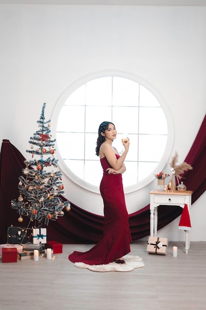 Portrait of a pretty young girl wearing a red gown and drinking wine smiling at the camera standing in decorated Christmas living room indoors