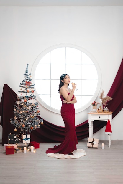 Portrait of a pretty young girl wearing a red gown and drinking wine smiling at the camera standing in decorated Christmas living room indoors