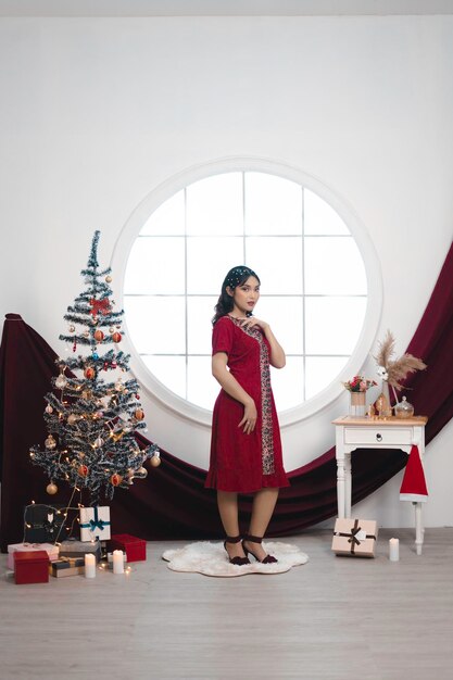 Portrait of a pretty young girl wearing a red gown smiling at the camera standing in decorated Christmas living room indoors