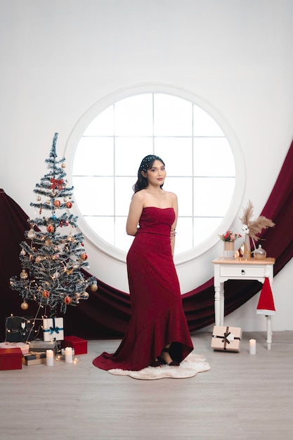 Portrait of a pretty young girl wearing a red gown smiling at the camera standing in decorated Christmas living room indoors